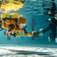 A humanoid robot and a person underwater in a pool. The robot is holding a plastic rod in two hands.